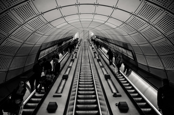 The image presents a striking black and white view of a modern subway station interior, focusing on the escalators. The composition is symmetrical, with two parallel escalators ascending diagonally towards the top center. The scene is dominated by the repeating patterns of the escalator steps and the architectural details of the tunnel.

The tunnel's ceiling is arched and lined with large panels, each punctuated with a grid of small, evenly spaced holes. This design creates a textured effect and adds visual interest. The walls are smooth and reflective, adding to the sense of depth and modernity. The lighting is diffused and even, casting soft shadows and highlighting the clean lines of the architecture.