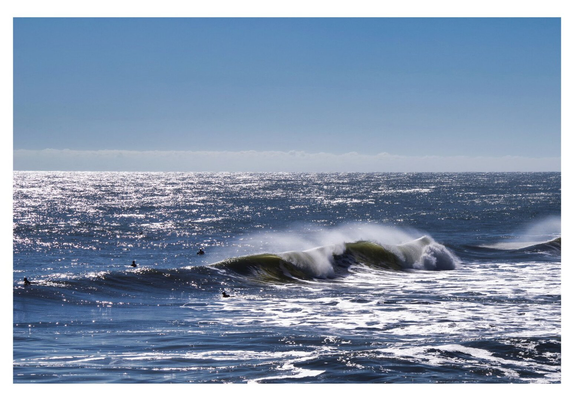 Surfers ride powerful, crashing waves in the ocean under a bright blue sky. The sun reflects off the water, creating a shimmering, energetic scene.