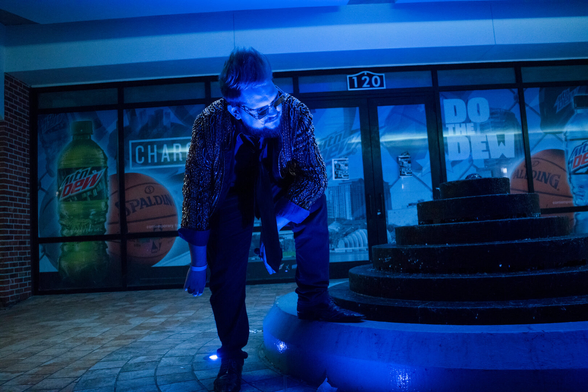A person in a sparkled coat stands near a water fountain at Downtown Orlando. A window advertisement is behind the scene.