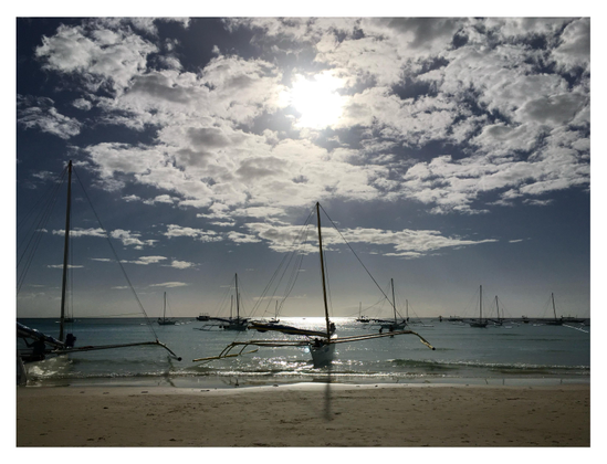 Photo taken on an island beach, looking out to sea. Several small sailboats, with long outriggers extending from their sides, float offshore with their sails furled. The central boat casts the shadow of its mast onto the sand in the foreground. The midafternoon sun at top center frame shines through some clouds and casts glittering light on the waves.