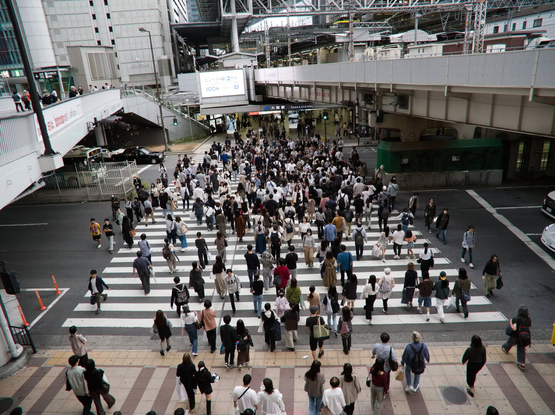 A photo of a wide zebra crossing between two pedestrian bridges crossing the same street. There is a dense crowd of people crossing the street in both directions. In the background there is sophisticated construction of business centers near the Osaka-Umeda station.