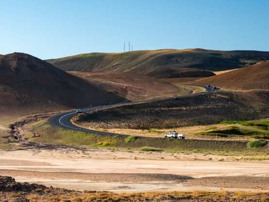A two-way asphalt road going up between brown/green hills. A few vehicles are driving on it, including a large white mountain car with a small trailer. The foreground is light-colored dirt.