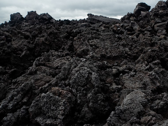 Black lava rock covering the vast majority of the image, with rugged texture. On top of the image there's a bit of grey menacing sky with clouds.