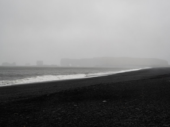 A very gloomy landscape with black sand in the foreground, grey sea (with very light hints of green), and in the background rocks protruding from the sea, barely visible despite their large size because of thick fog.