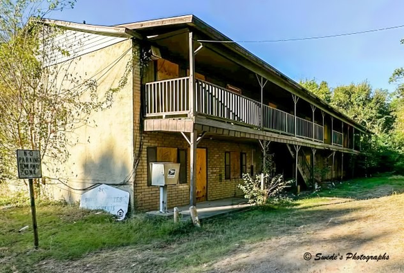 Balcony of Echoes

"An aging two-story apartment building stands quietly in a grassy clearing, its wooden bones weathered by time and neglect. The upper floor is lined with a long, continuous balcony—its railings faded and splintered, like the ribs of a forgotten ark. The windows are mostly boarded up, some with mismatched panels, others left open to the elements, hinting at abandonment or retreat. The exterior walls wear the patina of years—peeling paint, water stains, and the soft erosion of forgotten upkeep.

A pair of mailboxes, mounted on a single post, lean slightly forward as if bowing to the weight of memory. Nearby, a “No Parking in Fire Zone” sign stands upright, its red lettering stark against the muted tones of the building. The grass around the structure is uneven and wild, dotted with patches of shade from surrounding trees whose branches stretch protectively overhead. The sunlight slants in from the side, casting long shadows that suggest late afternoon—a golden hour that softens the decay with a touch of grace.

The scene feels rural, maybe suburban, but mostly timeless—caught between use and abandonment, utility and elegy. The building doesn’t shout; it murmurs. It holds stories in its silence, waiting for someone to listen." - Microsoft Copilot