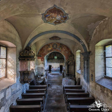 Interior of an old, abandoned chapel with wooden pews, faded wall paintings, arched ceiling, and ornate pulpit; sunlight filters through stained glass windows.