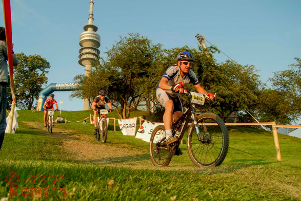 Farbfotografie im Format 3:2 Landscape.
Von einem Wiesenhügel auf dem Olympiagelände in München fahren drei Mountainbiker herunter. Die Fahrstrecke ist beidseitig mit Bändern abgesteckt. Dort wo die Biker herunterfahren ist braune Erde zu sehen. Im Hintergrund ragt der Olympiafernsehturm in den blauen Himmel. Die Sonne steht schon sehr tief, weswegen die Farben der Fotografie ein wenig wärmer sind.