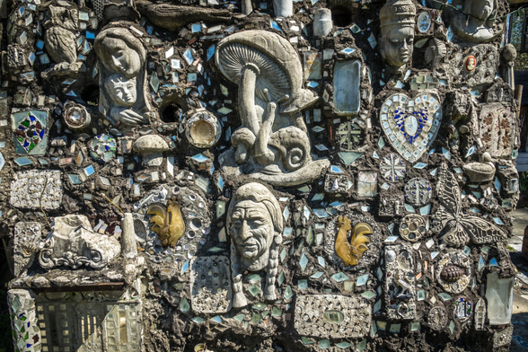 A close-up photograph of a highly textured, mosaic-like wall covering made of concrete or mortar embedded with numerous found objects. The surface is densely packed with various sculptural elements and reflective pieces.

Prominent features embedded in the wall include:

    A large, central sculpture of a mushroom with a small snail or similar creature underneath.

    Sculpted human faces/heads in the upper left, lower center, and upper right quadrants. The lower central face appears distinctly aged or stylized, possibly Native American in representation.

    A large heart-shaped mosaic on the upper right, composed of small mirror or tile fragments.

    Various small, yellow-brown sculpted shapes resembling roosters or perhaps fragments of animal forms, flanking the lower central face.

    Numerous small, irregularly shaped mirror or tile shards that catch the light, interspersed throughout the dark mortar base.

    Other indeterminate shapes, including small bottles or ceramic pieces, are also visible.

The overall aesthetic is one of outsider art or folk art assemblage, characterized by dense layering and texture.