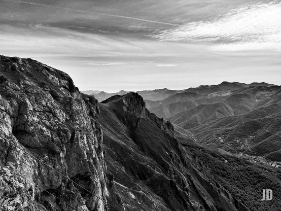 Fotografía en blanco y negro de un vasto paisaje montañoso. En primer plano, a la izquierda, una imponente ladera rocosa y empinada con texturas ásperas y cables diagonales que descienden. La imagen se abre hacia una serie de valles y cadenas montañosas ondulantes cubiertas de densa vegetación. En uno de los valles más profundos, se distingue un pequeño asentamiento o pueblo. Al fondo, las montañas se desvanecen en la distancia bajo un cielo con nubes dispersas y una estela de avión. La vista es desde una gran altura, capturando la inmensidad del terreno.