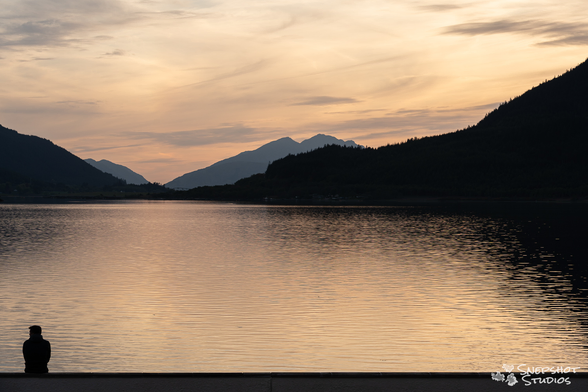 a photo of a Scottish Loch close to sunset. A person is sitting on a wall in the bottom left corner looking out over the water. They are only visible as a silhouette.