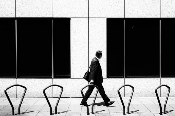 A businessman in a dark suit walks past a spotless, bright facade. The sidewalk appears just as spotless and clean. Only on the dark windowpane of a shop window is there a white bird dropping, which catches the businessman's eye. The bird dropping destroys the perfect world through which the perfect businessman moves.