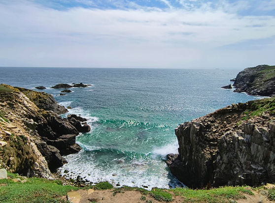 Von der Insel oben fotografiertes türkis blaues Meer, mit einer Welle kurz vor dem Auftreffen auf die Insel, eingerahmt von Felsenklippen. Der Horizont ist bewölkt, am oberen Bildrand sieht man das blau des Himmels.