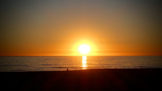 Wie eine leuchtende Apfelsine liegt die Sonne auf dem Meer, kurz davor hinter den Horizont zu tauchen. Der Himmel ist gefärbt in Orange, Gelb und Ockertönen. Der Strand im Vordergrund ist nur noch ein dunkler Schatten. Dazwischen glänzt das ruhige Meer in mattem Silberschwarz und reflektiert einen schmalen gelben Sonnenstrahl. 