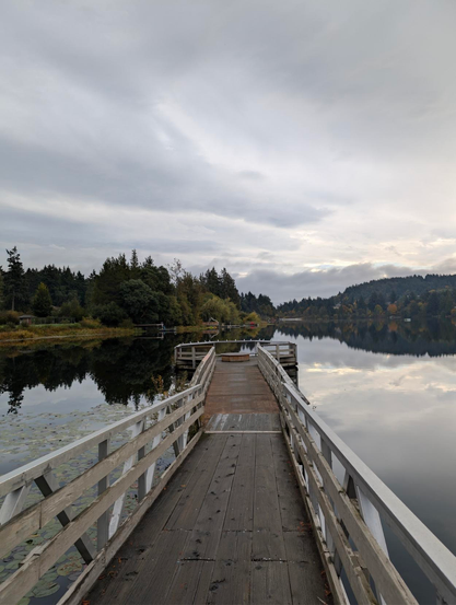 Looking down a short, small fishing pier? Dock? At a flat Glen lake. The sky is heavily overcast, some of the leaves on the trees surrounding the lake are changing colour.