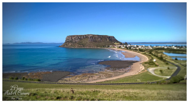 A wide angle, tilt shift style photograph showing a panoramic view of The Nut (a flat-topped, ancient volcanic plug) and the surrounding coastline in Stanley, Tasmania.

The sheer, grassy cliffs of The Nut dominate the center, overlooking a crescent shaped sandy beach, turquoise ocean water, and a small town. The foreground is a grassy hill leading down toward the beach road.
