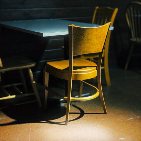 A wooden interior chair sits against a table, cast under a warm-hued spotlight in a darkened room.