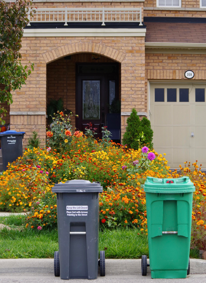 A Home with a flower garden on the front lawn and some Waste Management bins near the house and curb-side.