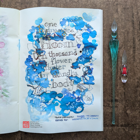 Photo of notebook next to two glass dip pens, one of which is just a tip. The page shows a handwritten haiku surrounded by ink splotches in varying shades of vivid blue resembling an explosion of flower petals. Some of the edges of the ink splotches are tinged with crimson, and bits of the lettering overlapping the splotches gleam with gold. 
"one hydrangea bloom
ten thousand flower cluster
my skandha body"
- Poetry and art by Maiji/Mary Huang, humangray.com