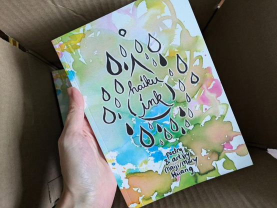 Hand holding the haiku ink book, showing off the cover with colourful ink splashes and ink brush hand-written cover text with many illustrated drops of ink raining down: "haiku ink - poetry & art by Maiji/Mary Huang".