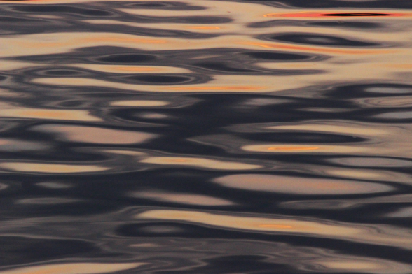 Photograph of black, grey, and orange shapes of light reflected on a lake surface, forming abstract ovals and swirls.