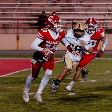 Three football players from bottom left to upper right all leaning to the right with mostly empty bleachers behind. In front Red #18 holds the football, white #55 is running after him with a red player following