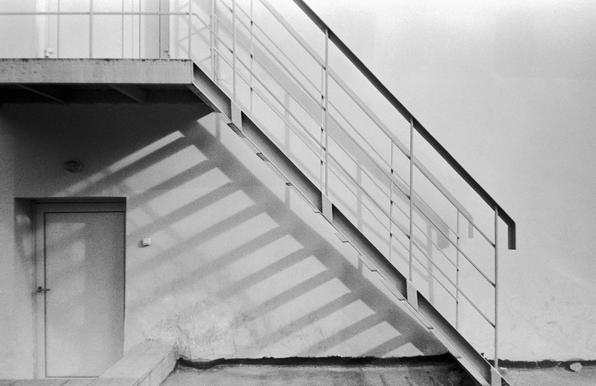 In the black-and-white photo, the staircase is shown from the side. The shadows of its railings fall on the building's wall.