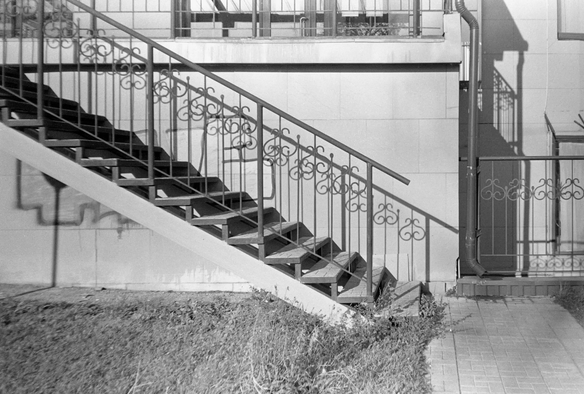 In the black-and-white photo, the staircase is shown from the side. The shadows of its railings fall on the building's wall.