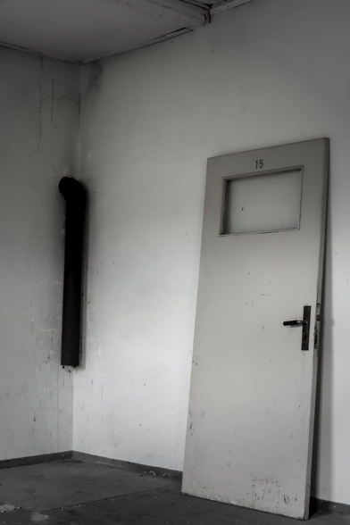 Aufnahme einer schräg an die Wand gelehnten Tür mit der Nummer 15 in der Lagergedenkstätte Sandbostel, daneben ein schwarzes Rohr an der Wand.