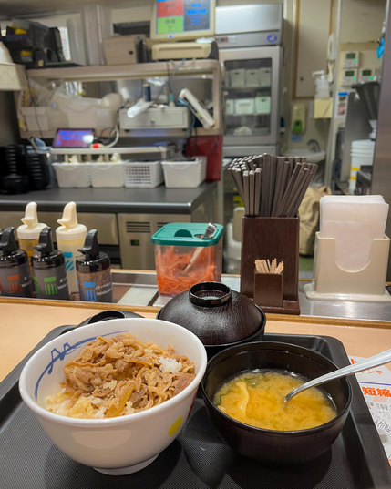 Una bandeja con un bol de gyudon (ternera a la plancha cortada muy fina sobre arroz blanco) y una sopa miso. De fondo, la cocina (abierta) del local.