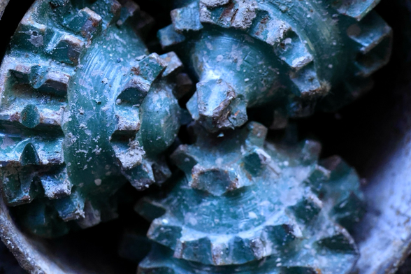 Drei kegelförmige Bohrer sitzen in einem Rohr und ergeben zusammen einen Bohrkopf. Die Bohrer sind im mittleren Bereich mit zwei Zahnreihen und am oberen Bereich mit einer Zahnreihe umgeben. Die Bohrer schimmern leicht grün. Die Spitzen der Kegel treffen sich in der Mitte. 