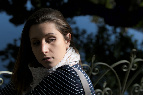 A photo of a young woman with mid long brown hair, wearing a blue top with white thin horizontal stripes, and a white scarf with floral pattern. She’s looking backward above her left shoulder. The right side of her face is in the shadow and the left side is lit by the sun, creating a big contrast
