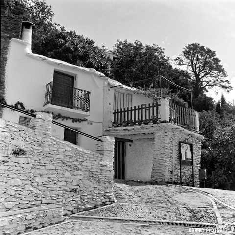 Two white stone houses with balconies and railings line a cobblestone street on a sloping terrain; this charming scene, captured in black and white on film, features trees and greenery in the background under a partly cloudy sky.