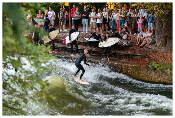 Mensch in schwarzem Neopren surft auf einer Welle in einem Bach. Am Ufer stehen weitere Surfer und Schaulustige.