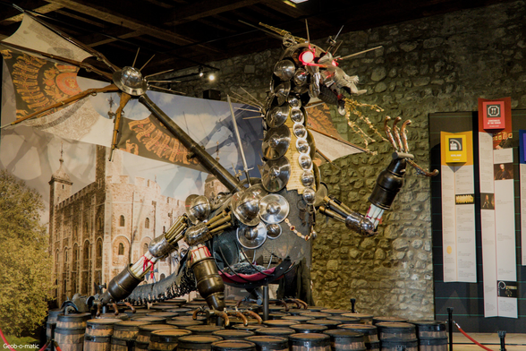 Photo of a metallic sculpture made of many old british soldier helmets, armour pieces and chain-mails, rifles… to form a dragon with red glowing eyes
