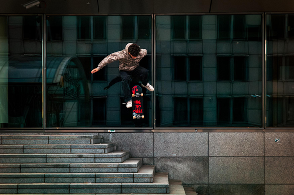 The image captures a skateboarder mid-air, performing a trick against an urban backdrop. The primary subject is the skateboarder, who is suspended in the air above a set of concrete stairs. The skateboarder is wearing a hooded jacket with a camouflage pattern, dark pants, and white sneakers. His body is contorted in a dynamic pose, suggesting the execution of a skateboarding maneuver. The skateboard is prominently displayed beneath him, featuring a black deck with red and white graphics.

The background is dominated by a large glass facade that reflects the surrounding architecture. The reflection reveals a building with numerous windows, creating a sense of depth and urban environment. The glass facade has a dark, slightly greenish tint, contributing to the overall cool color palette of the image.
