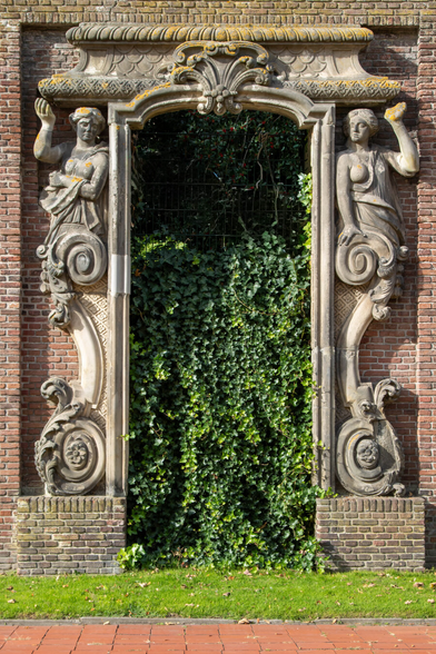 A rectangular gate is set into a wall of small brown-red stones, surrounded by a border of stone ornaments. At the top, on either side, stands a Greek-style sculpted, busty woman. Together, they support a stone roof above the door. The doorway is closed with wire mesh, completely overgrown with ivy.