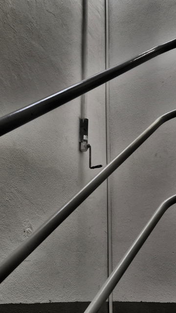 Ein Schwarzweißfoto, das in einem Treppenhaus eines Industriebaus aufgenommen wurde. Im Zentrum des Bildes ist an einer Wand eine Handkurbel zu sehen, von der oben ein Verbindungsstab aus Metall aus dem Bild läuft. Vor der Kurbel verlaufen drei Rundrohre aus Metall von einem Treppengeländer von unten links nach oben rechts durch das Bild.