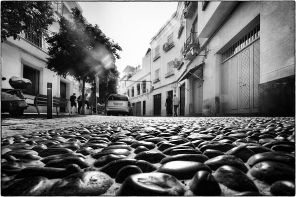 Eine enge, von weissen Gebäuden gesäumte Gasse in einer südländischen Stadt. Der Boden ist mit unregelmässigen, runden Kopfsteinpflastersteinen bedeckt, die im Vordergrund stark hervortreten. Links steht ein Motorroller, rechts sind geschlossene Garagentore zu sehen. Einige Menschen gehen in der Gasse, die von Bäumen und Laternen gesäumt wird. Im Hintergrund erkennt man ein geparktes Auto. Die Aufnahme ist in Schwarz-Weiss gehalten und wirkt sehr kontrastreich.