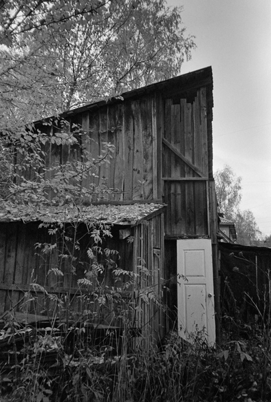 The photograph depicts an old wooden building, a barn, surrounded by vegetation. The barn walls appear weathered, bearing the marks of time and the elements. The barn has a white door that contrasts with the dark background of the walls. The area around the building is overgrown with tall grass and shrubs, creating a sense of abandonment and isolation. The cloudy sky in the background enhances the melancholy atmosphere of the photograph.