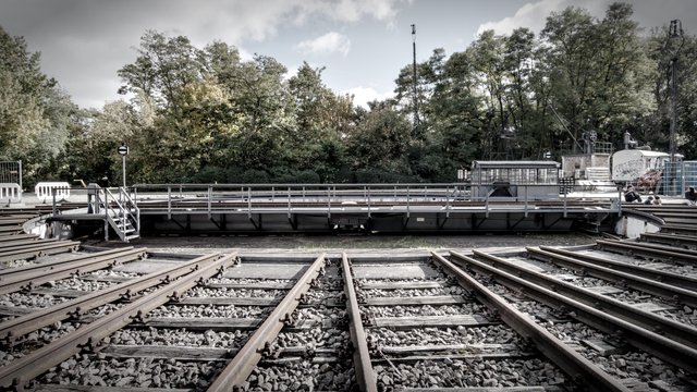 Side view of a railway turntable. The foreground is dominated by railroad tracks going concentrically into the round pit, in the background the rotating element can be seen going from left to right, with a stair leading into the pit on the left side. In the background, there are trees and a slightly clouded sky.