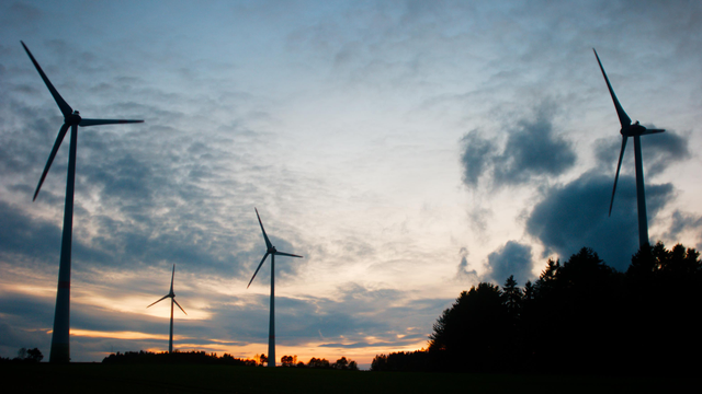 Sunset picture. Four wind turbines sticking into the sky as black silhouettes above a black forest outline, with the sky colored salmon and light blue, half covered by dark lead-colored clouds.