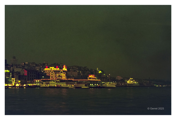 Nighttime harbor scene with neon-lit buildings reflecting on the water. A ferry is docked, and city lights illuminate a hilly coastal area, creating a calm, vibrant atmosphere.