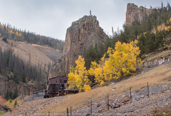 Rusty metal, multi-story mine building sets on a slope below a dominant rock pinnacle next to a grove of golden aspen. A ridge in the background is covered in mine tailings. Flat grey sky.
©BosqueBill.com