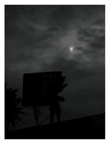A bright, thin crescent of the moon is visible through a dark, cloudy sky. In the foreground, the silhouettes of palm trees and a basketball hoop stand in stark contrast against the eerie, dim light.