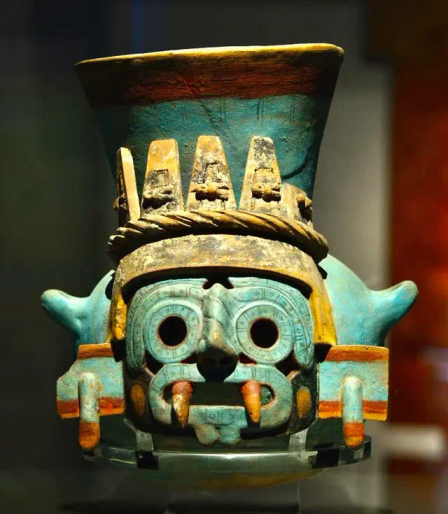 a turquoise blue pottery sculpture, the top a wide column angled outwards like a vase, the face is a fearsome one with large red fangs, large circular black eyes, it has a gold crown on, it is looking directly at the viewer 