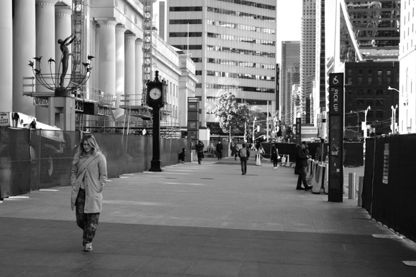 On Front Street outside the station, we see the towers of downtown in the distance. In front of the station’s columns are the freestanding clock and a wrought iron sculpture of a figure reaching to the sky. Along the street are barriers to ongoing construction in various spots. We see pedestrians in the distance and waiting on the edge of the street. In the lower left corner of the image, a woman in a long coat walks toward the camera, her head looking down.