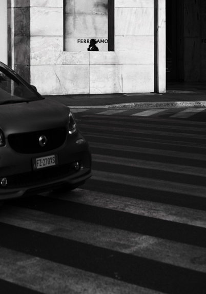 A black and white image of a car approaching a pedestrian crossing, while the silhouette of a man on the phone is partly visible and in-focus on the opposite side of the road, as he passes behind a white marble wall with a large rectangular opening in it.
