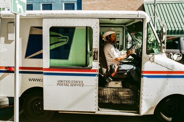 A photo of a mail carrier in hist United States Postal Service delivery van.  He is sitting in the drivers seat and wearing a white 'cowboy' type hat.