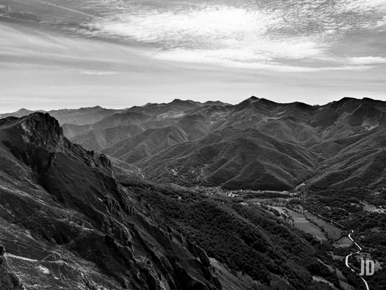 Una vista panorámica en blanco y negro de un vasto paisaje montañoso. En primer plano a la izquierda, una montaña rocosa y escarpada se eleva con laderas empinadas y acantilados pronunciados. Hacia el centro y la derecha, un valle profundo y sinuoso se extiende, flanqueado por numerosas montañas cubiertas de densa vegetación y árboles. En el fondo del valle, se vislumbra un pequeño pueblo o caserío, con campos y un río serpenteante que lo atraviesa. En la distancia, capas de montañas más pálidas se desvanecen hacia el horizonte, bajo un cielo parcialmente cubierto por nubes tenues y alargadas. La fotografía en blanco y negro acentúa los contrastes de luz y sombra, dando a la escena una atmósfera dramática y atemporal.