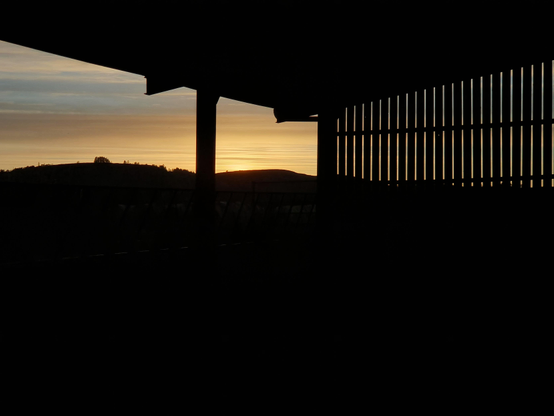 Looking out from a farm steading at a sunset sky, streaked pale orange and blues.The silhouetted roof angles down from top left to a low point right of centre, the wall to the right has vertical slats which allows the light in